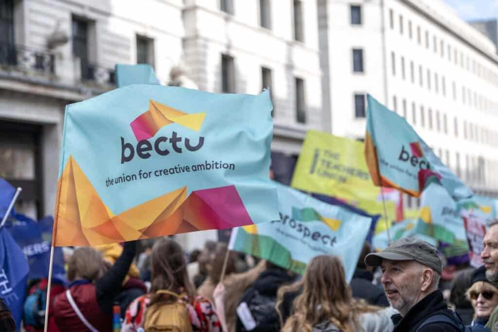 Group of protesters waving colorful flags with the 'bectu' logo and the slogan 'the union for creative ambition' on a city street edge.