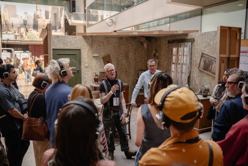 Man with glasses holding a microphone explains a vintage room setup to a crowd at an indoor exhibit. They wear headphones as part of the tour or presentation.