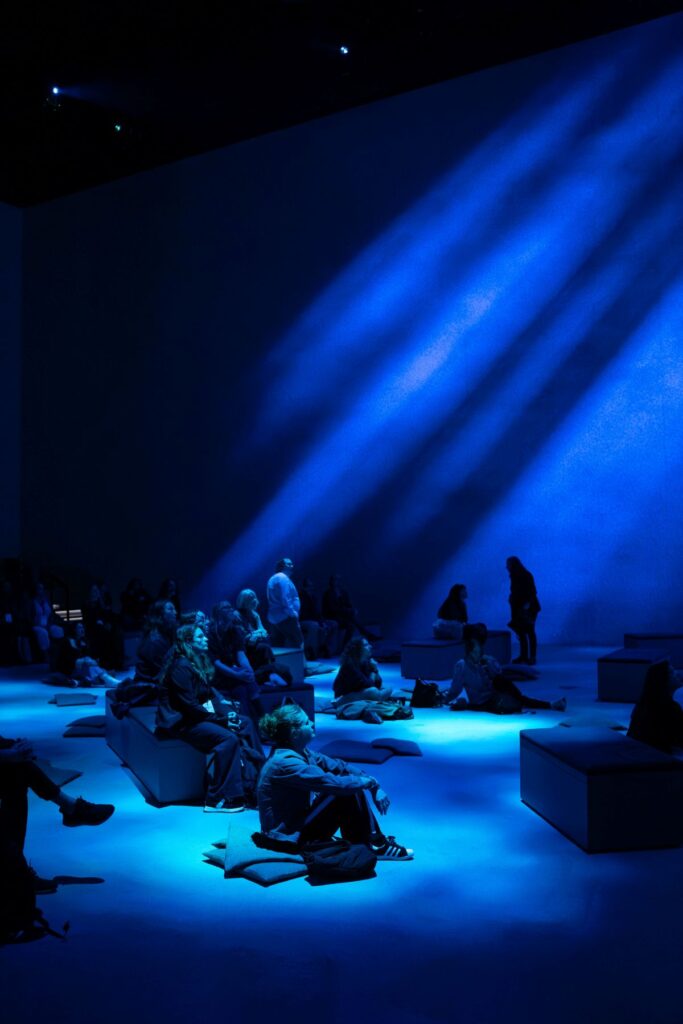 Audience seated on floor cushions in a dark auditorium swept by deep blue light beams.