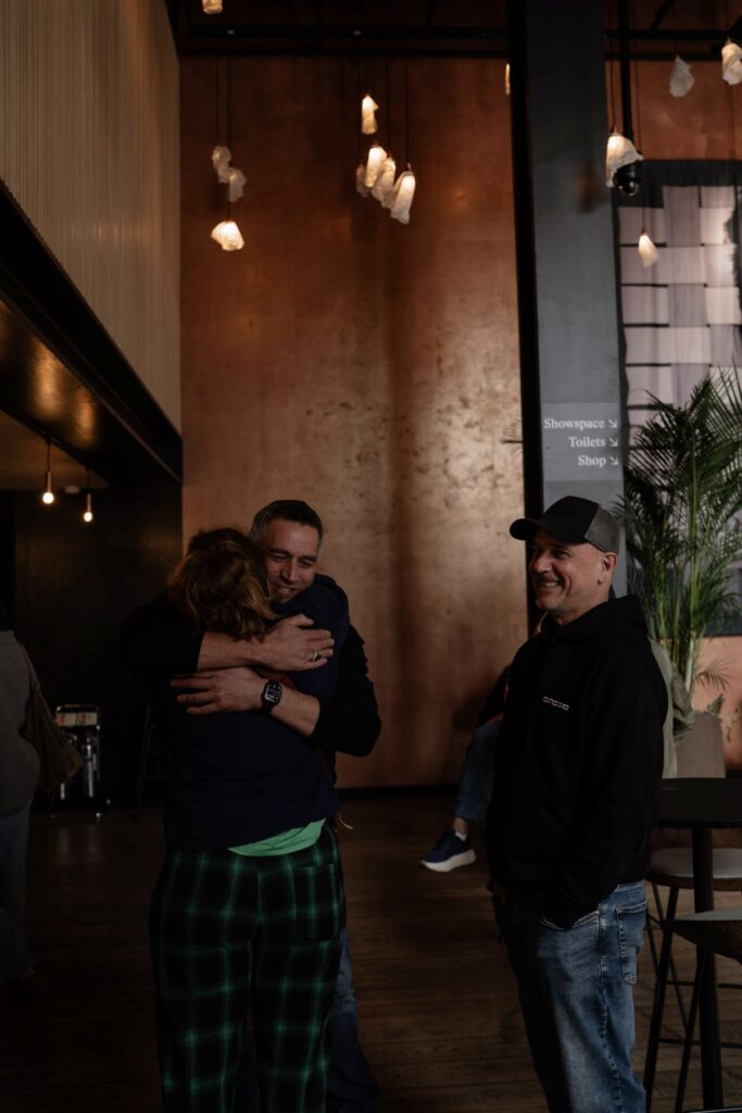 Two people hug in greeting in a dimly lit lobby while a man in a cap looks on smiling.