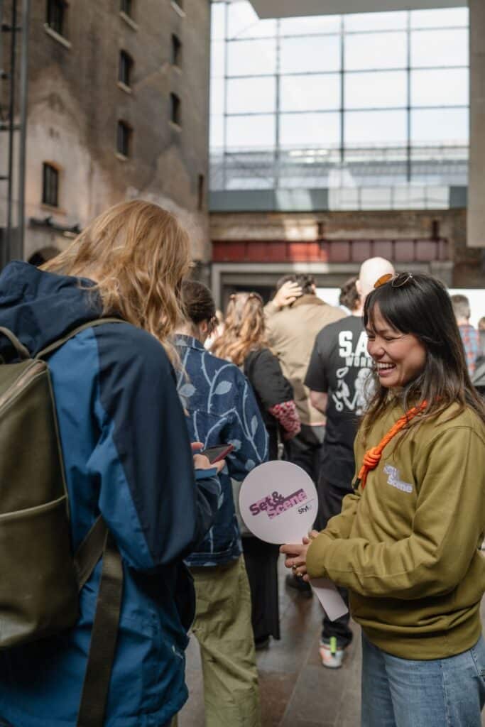 Attendees chat in a sunlit brick-and-glass atrium; a woman in an olive hoodie holds a circular Set & Scene hand fan.