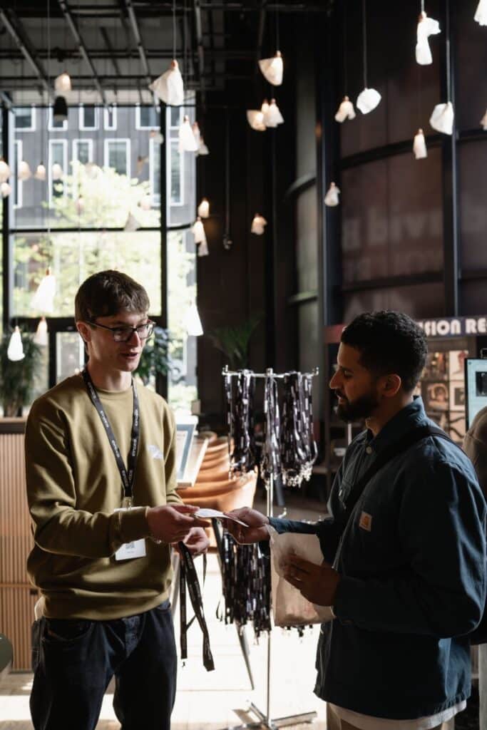 Two men wearing lanyards exchange a name badge in a dark showroom hung with sculptural pendant lights.