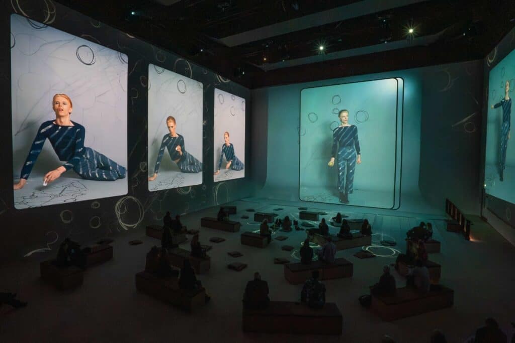 Audience on low wooden blocks watches an immersive gallery where large screens show a model in a blue patterned outfit.